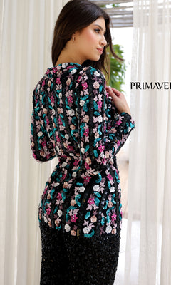A woman stands indoors facing away, wearing the Long Sequin Formal Jumpsuit: Primavera 4388, featuring colorful floral embellishments. Light curtains and greenery are visible in the background.