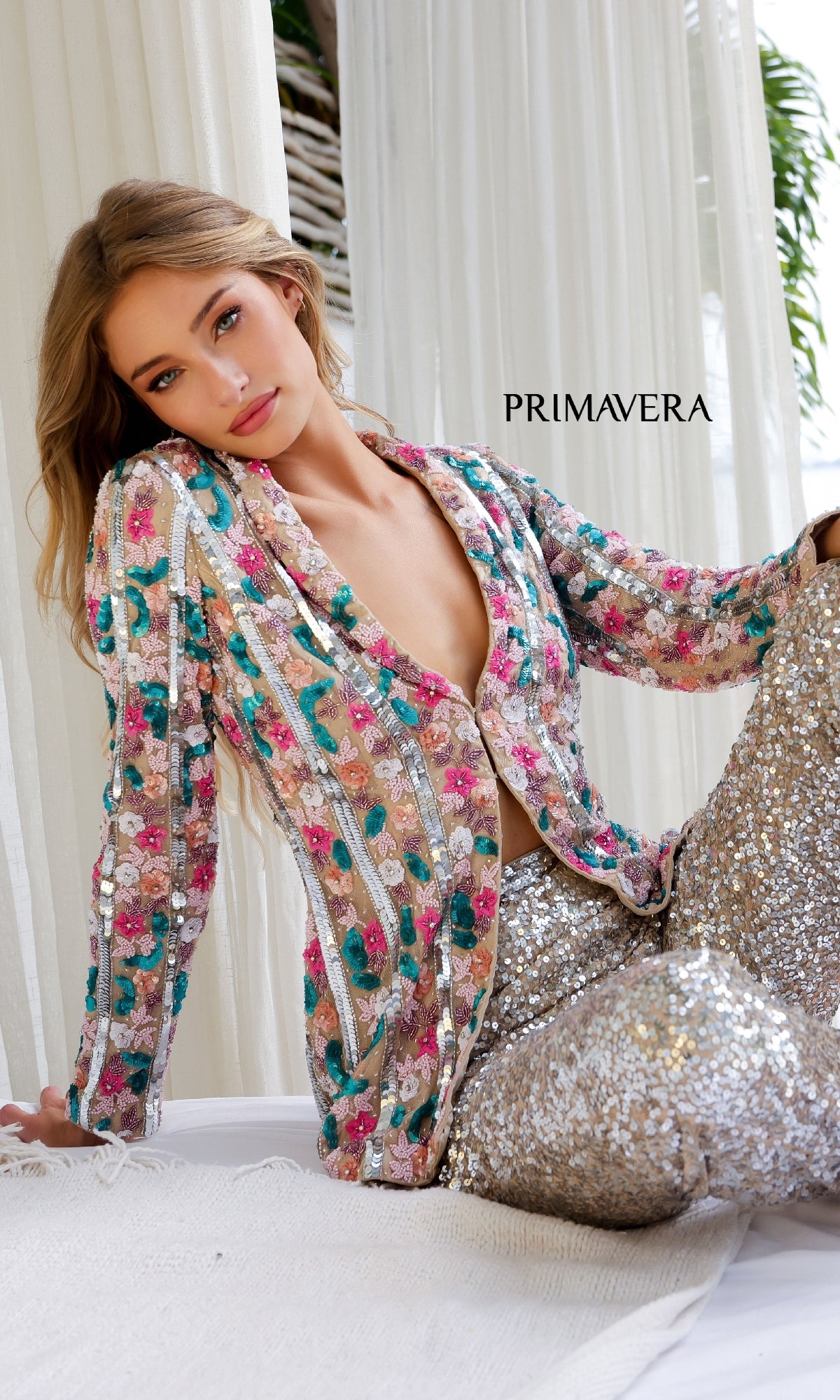 A woman wearing the Primavera 4388 Long Sequin Formal Jumpsuit sits on a white surface with white curtains and greenery behind her. The word 