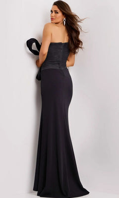 A woman stands facing away in the Jovani 43971 long prom dress—a black strapless gown with a pleated bodice and statement bow. She holds a black wrap, her long brown hair down, earrings gleaming, for an elegant formal look.