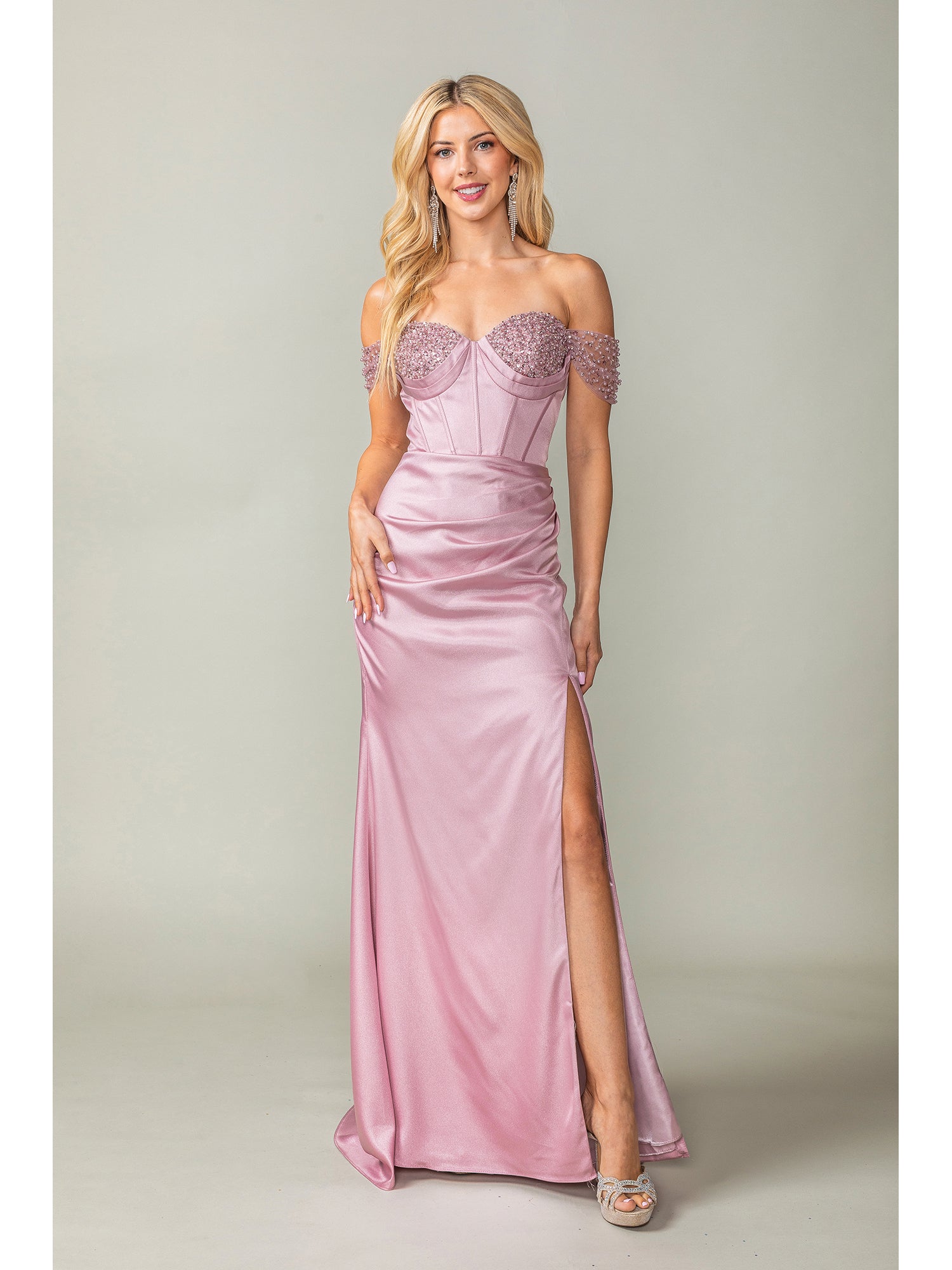 A woman wears the Off-the-Shoulder Long Prom Dress 4401 with beads, featuring a light pink hue, beaded straps, corset bodice, thigh-high slit, and silver heels against a plain background.