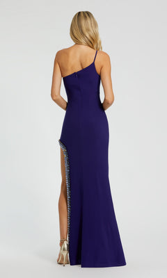 A woman stands facing away, wearing the Mac Duggal 44030 Bead-Trim Long Indigo Prom Dress with a one-shoulder design and high side slit, paired with metallic heels.