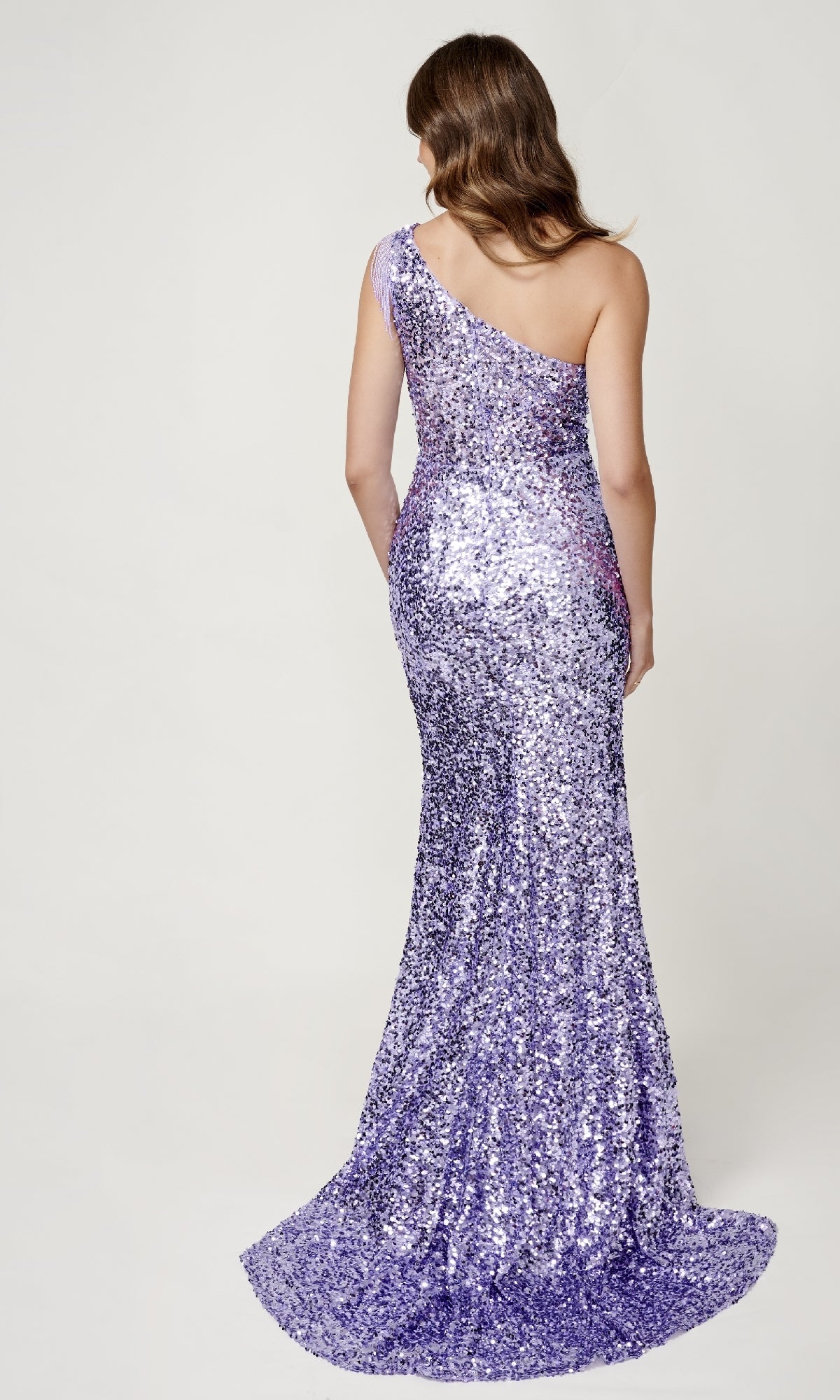 A woman with wavy brown hair models the Sequin Prom Dress: Nina Canacci 4405, featuring a one-shoulder neckline and floor-length purple sequins, shown from the back against a plain light background.