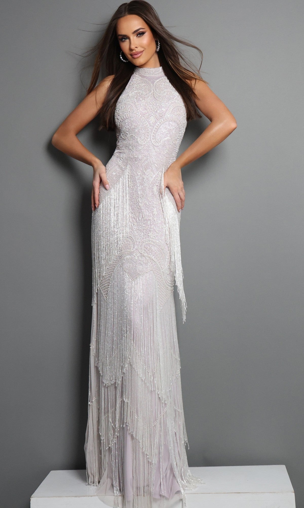 A woman stands against a gray background wearing the Jovani 44061, a sleeveless, floor-length beaded white formal dress featuring Art Deco fringe details.