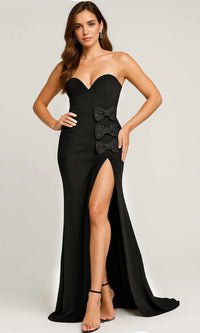 A woman wears the black Strapless Sweetheart Long Prom Gown Jovani 44066, featuring a thigh-high slit and waist bows, paired with black high-heeled sandals, as she stands elegantly against a plain background.