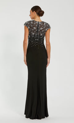 A woman, hair in a neat bun, faces away wearing the Mac Duggal 44081 Cap Sleeve Black Formal Dress—a floor-length gown with sheer embellished lace back and cap sleeves.