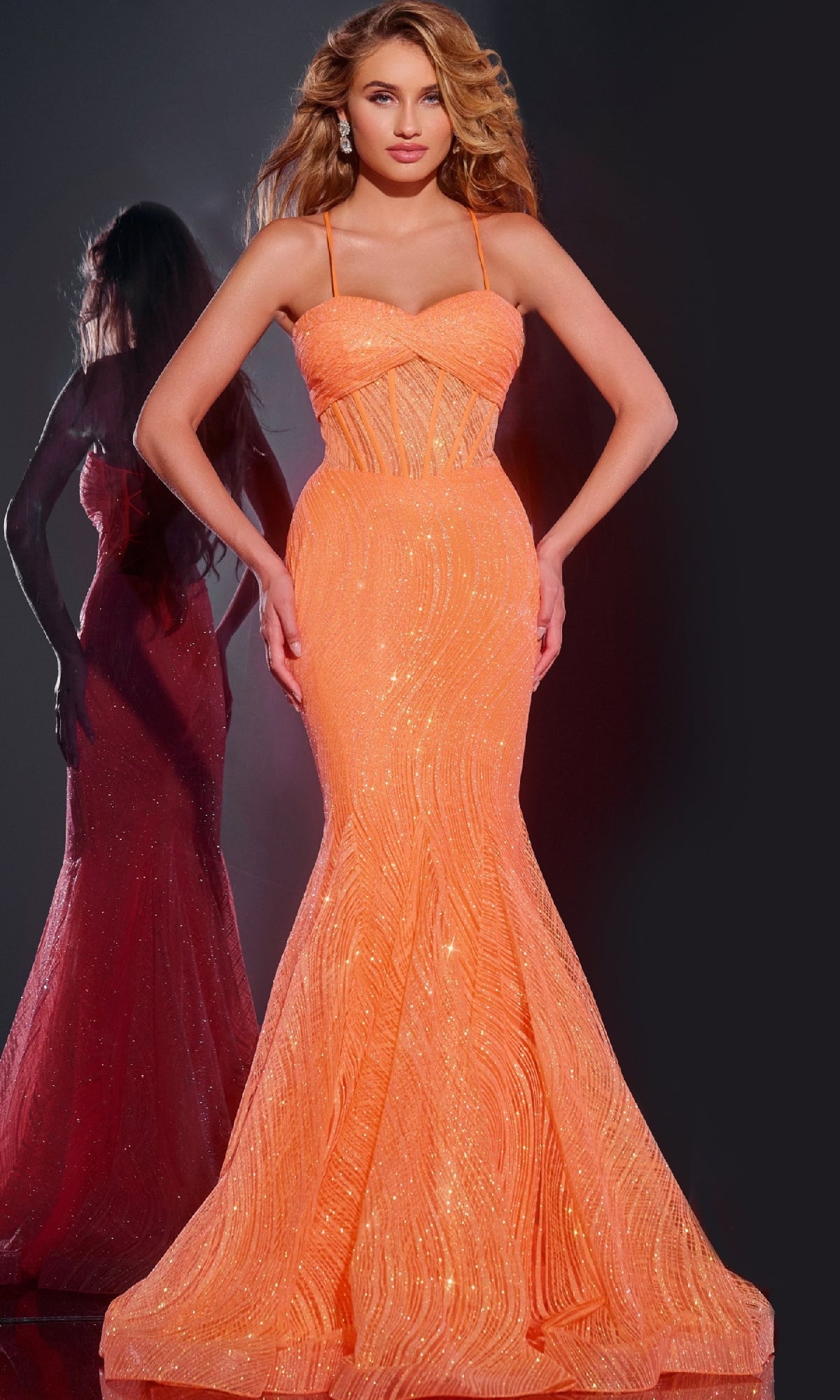 A woman poses in the Jovani 44208, a long orange mermaid prom dress with thin straps and shimmering sequins; her reflection appears behind her.