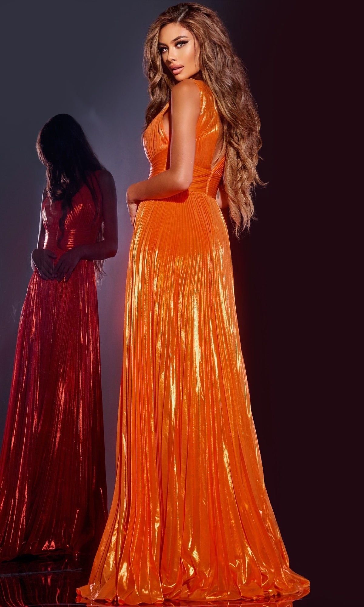 A woman stands before a dark background in the Jovani 44230 long prom dress, featuring an A-line skirt and bright orange pleats, with her reflection visible behind her.