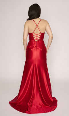 Woman wearing the Nina Canacci 4426 long formal dress in red satin, with a fitted bodice, v-neckline, criss-cross straps, lace-up back, and chic thigh-high slit, facing away from the camera.