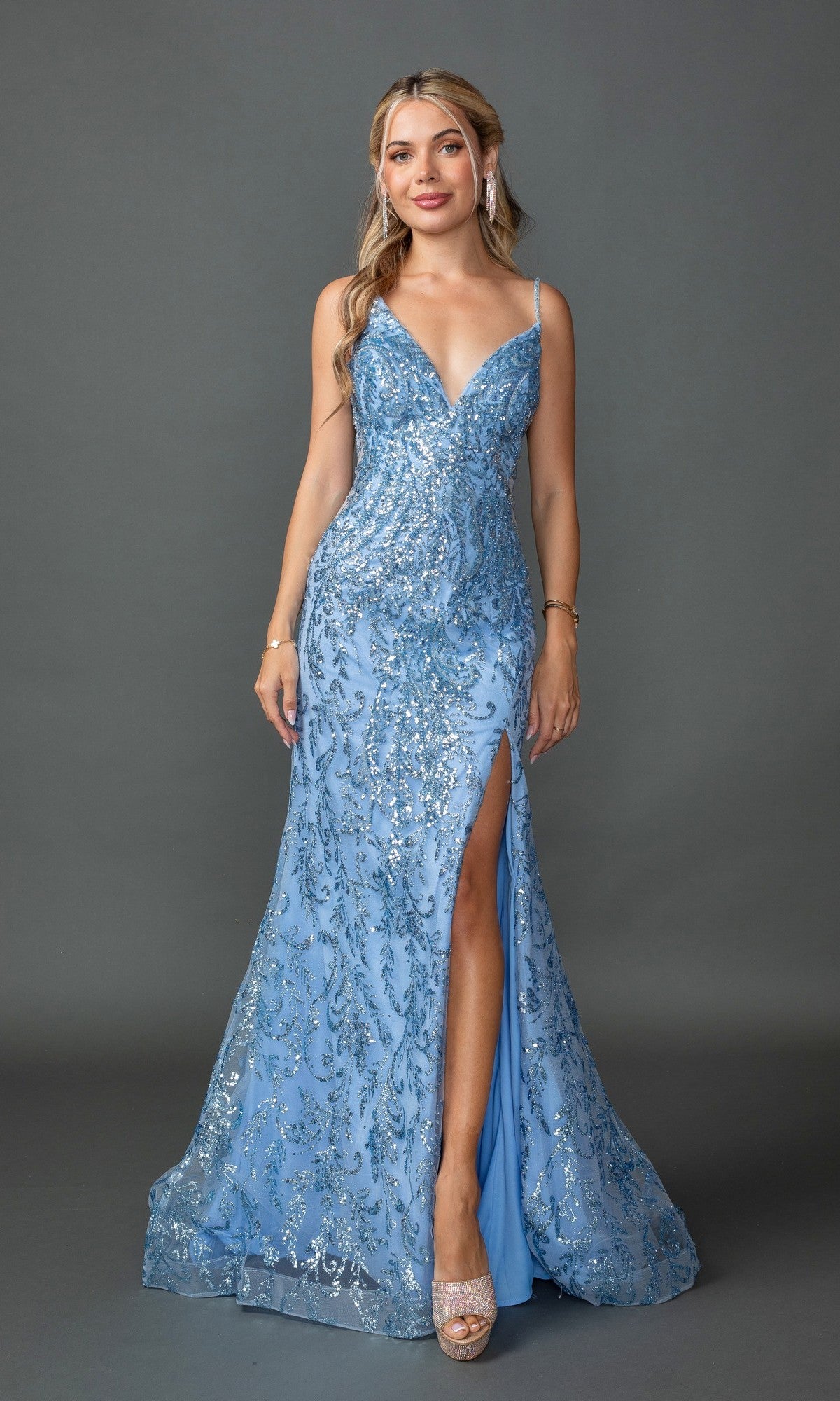 A woman models the Dancing Queen 4426 long sequin-print prom dress in Bahama Blue, featuring a floor-length silhouette with a thigh-high slit and lace-up open back, standing against a plain gray background.