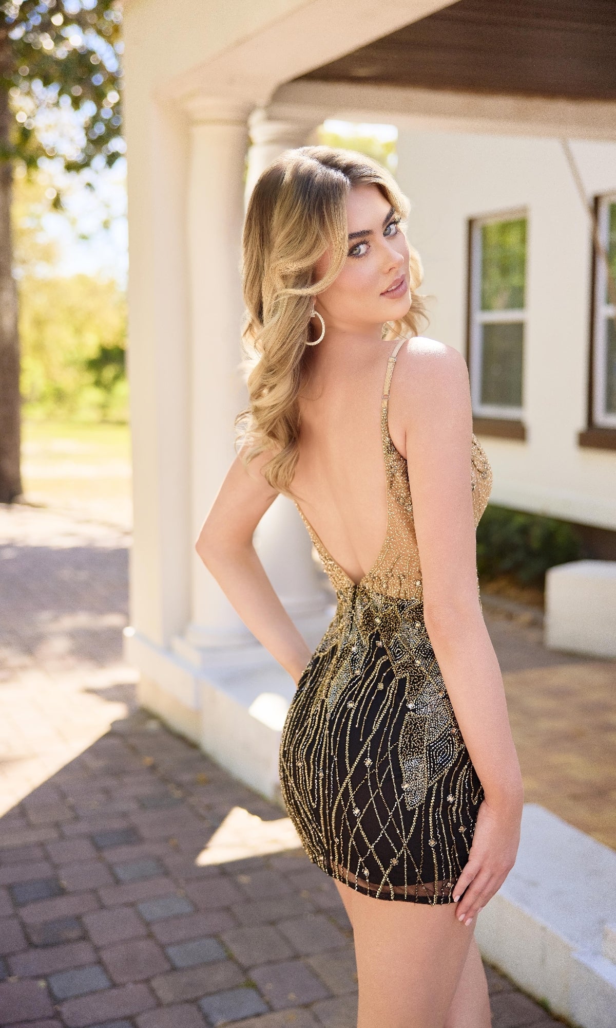 A woman with blonde hair stands outdoors on a paved walkway, looking over her shoulder and wearing the Primavera 4434 short homecoming dress in gold and black sequined embroidery with a fitted silhouette.
