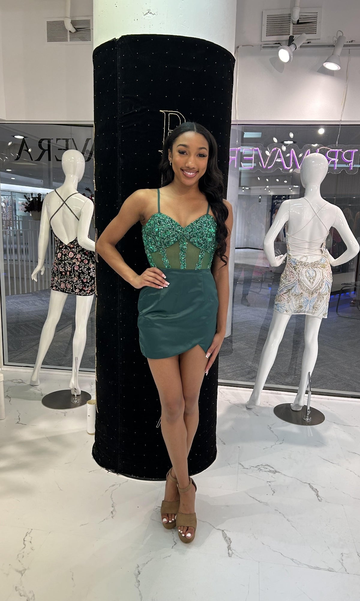 A woman poses indoors in front of two mannequins, wearing the Primavera 4437 short homecoming dress—a green sequined mini dress with a fitted satin wrap skirt—paired with tan heels.