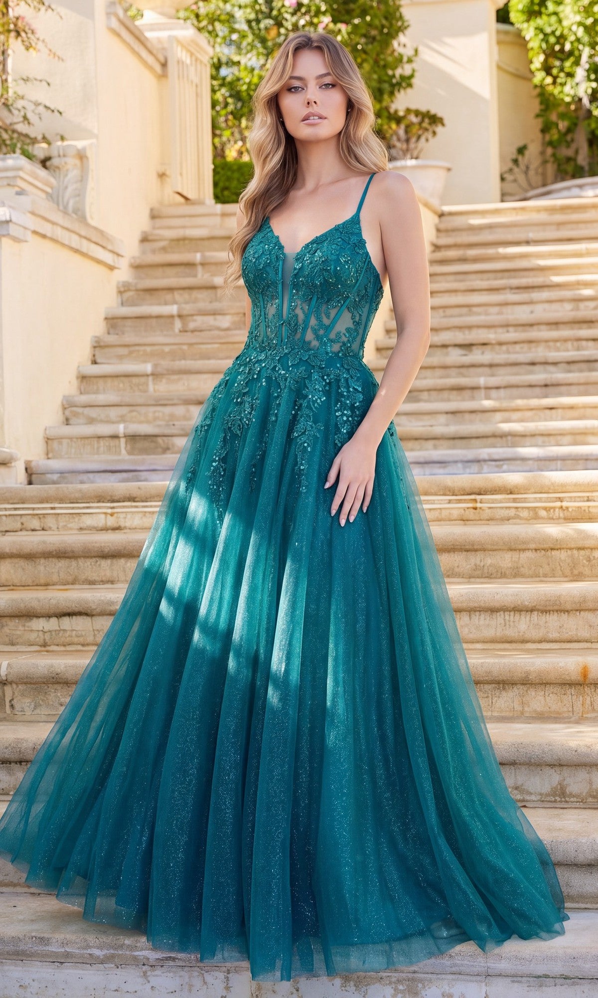 A person stands on stone steps wearing the Dancing Queen 4437 Long Tulle Prom Ball Gown in hunter green, featuring a sleeveless, lace-up fitted bodice and a flowing floor-length skirt. Lush greenery and part of a building are visible in the background.