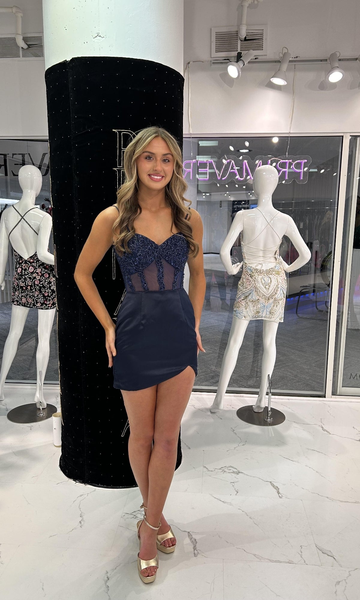 A woman stands indoors wearing the Primavera 4437 Short Homecoming Dress, which features a navy blue strapless design with a hot-stone embellished bodice and fitted satin wrap skirt. Two mannequins in decorative dresses are seen in the background.