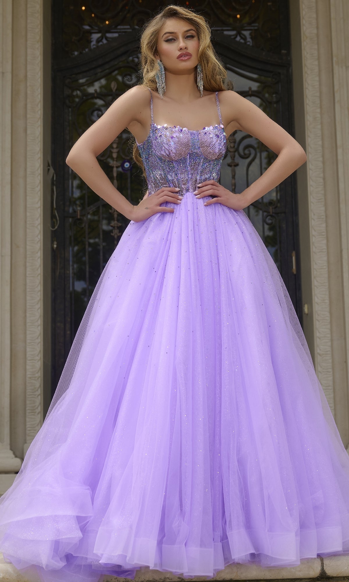 A woman stands on steps in front of ornate doors, wearing the lilac Beaded-Corset Long Prom Ball Gown Jovani 44511, featuring a fitted bodice and full shimmering tulle skirt.