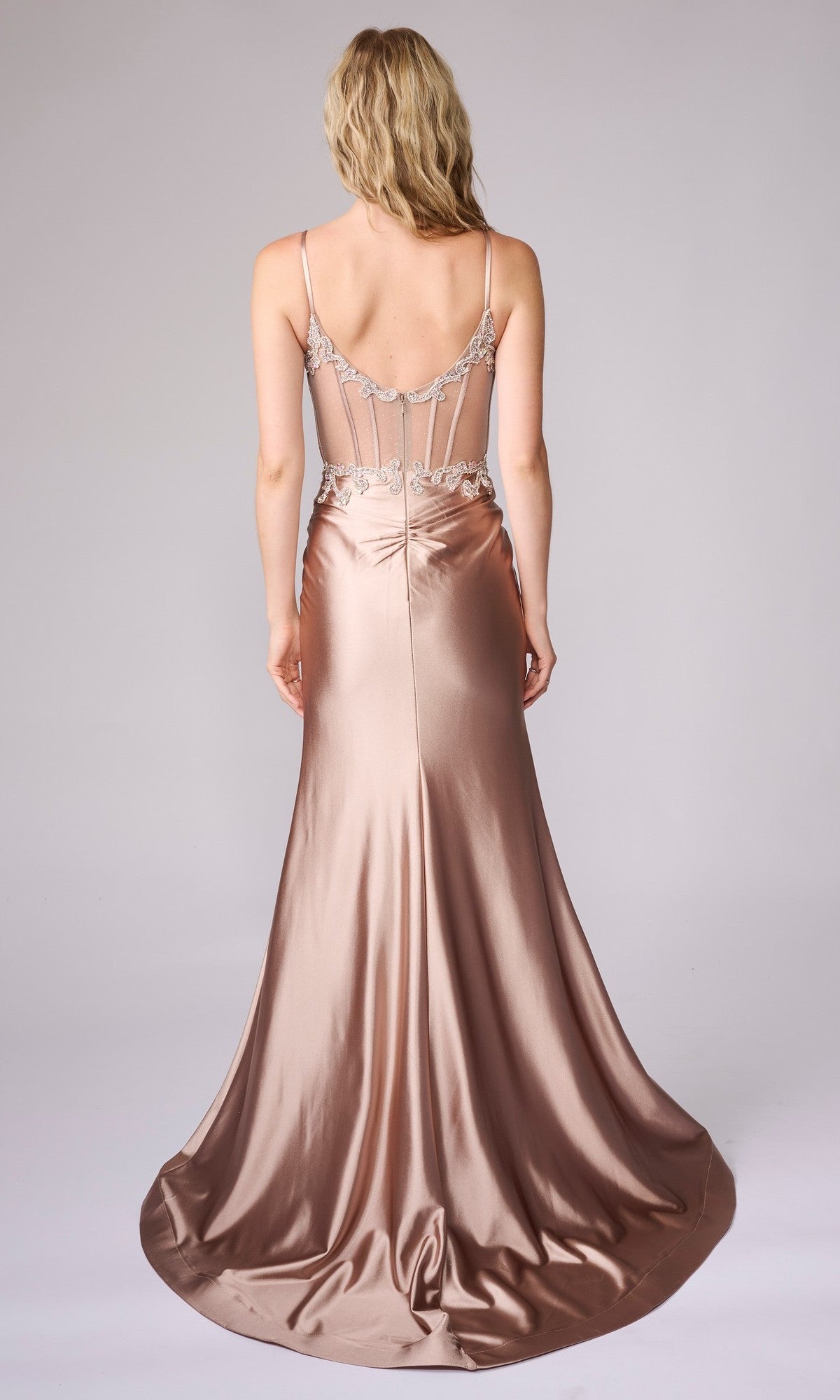 A woman faces away, wearing the Nina Canacci 4453 long formal dress in rose gold satin, featuring thin straps and an embellished sheer bodice.