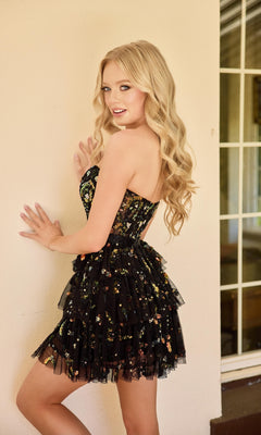 A young woman with long blonde hair poses indoors against a light-colored wall, wearing the Primavera 4463 short homecoming dress—a black, strapless fit-and-flare style with a floral pattern.
