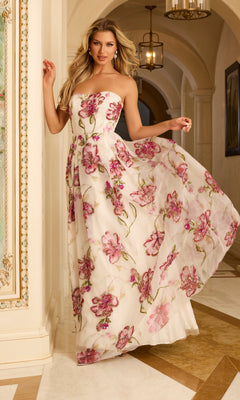 A woman wears the Long Prom Dress: Jovani 44645 in Off White/Multi, featuring a strapless floral ballgown design, as she stands in an ornate hallway holding her dress and smiling at the camera.