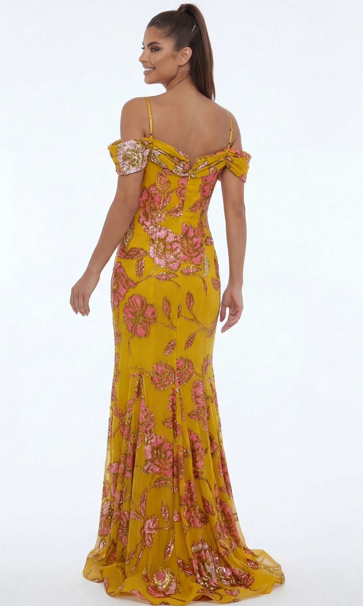 A woman stands facing away and smiling in a Jovani 44652 long formal dress, featuring an off-shoulder, floor-length silhouette with a yellow fabric adorned with pink floral patterns, set against a plain white background.