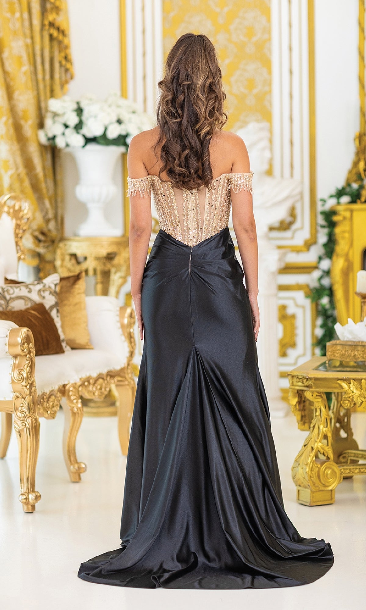 A woman with long wavy hair faces away in an ornate room, wearing the Sequin-Bodice Long Prom Dress: Dancing Queen 4486 with a beaded bodice and elegant off-the-shoulder sleeves.