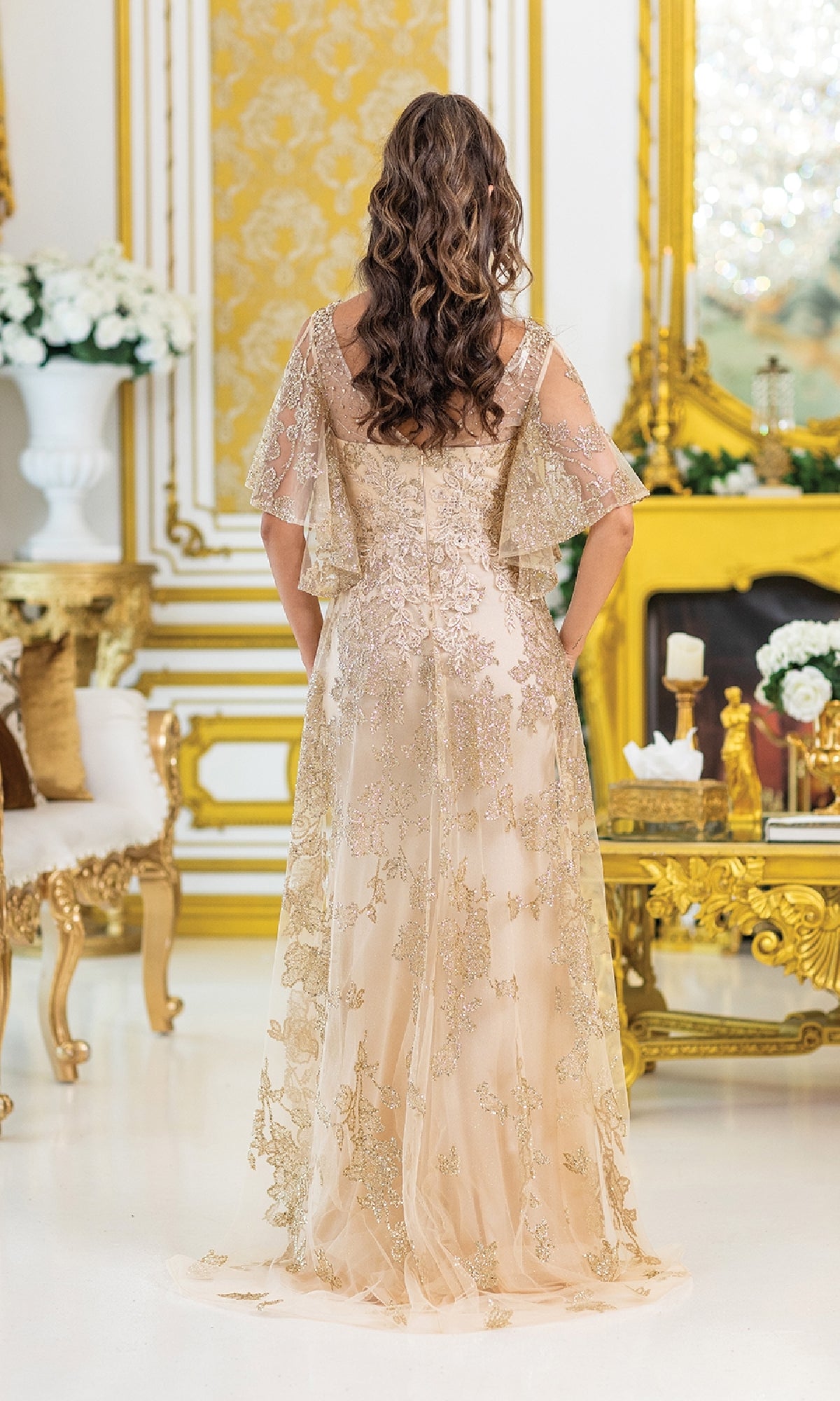 A woman with wavy brown hair stands with her back to the camera, wearing the Dancing Queen 4493 Removable Sleeves Prom Gown in an opulent gold and white room.