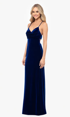 A woman stands smiling against a plain white background, wearing the Blondie Nites 4494BN dark blue prom dress—a floor-length, sleeveless velvet gown with thin straps and a graceful silhouette.