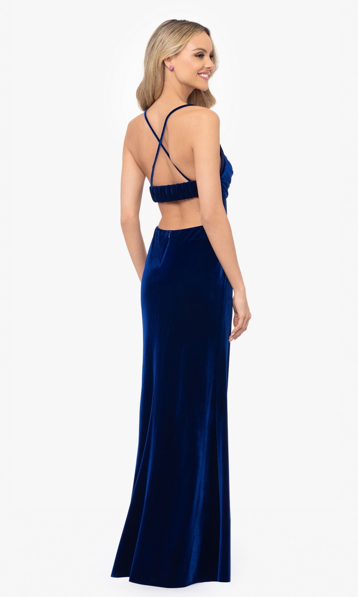 Woman in a floor-length, dark blue Blondie Nites 4494BN prom dress with thin crisscross straps and open back, standing and smiling while facing away from the camera.