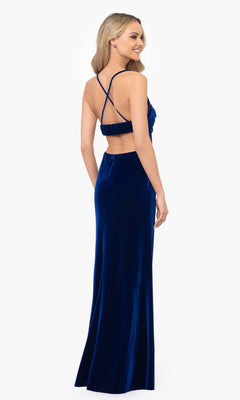 Woman in a floor-length, dark blue Blondie Nites 4494BN prom dress with thin crisscross straps and open back, standing and smiling while facing away from the camera.