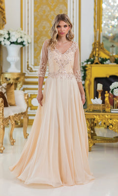 A woman wearing the Dancing Queen 4497 long sleeve formal dress with a beaded bodice and lace details stands in an ornate room adorned with gold accents and white flowers.