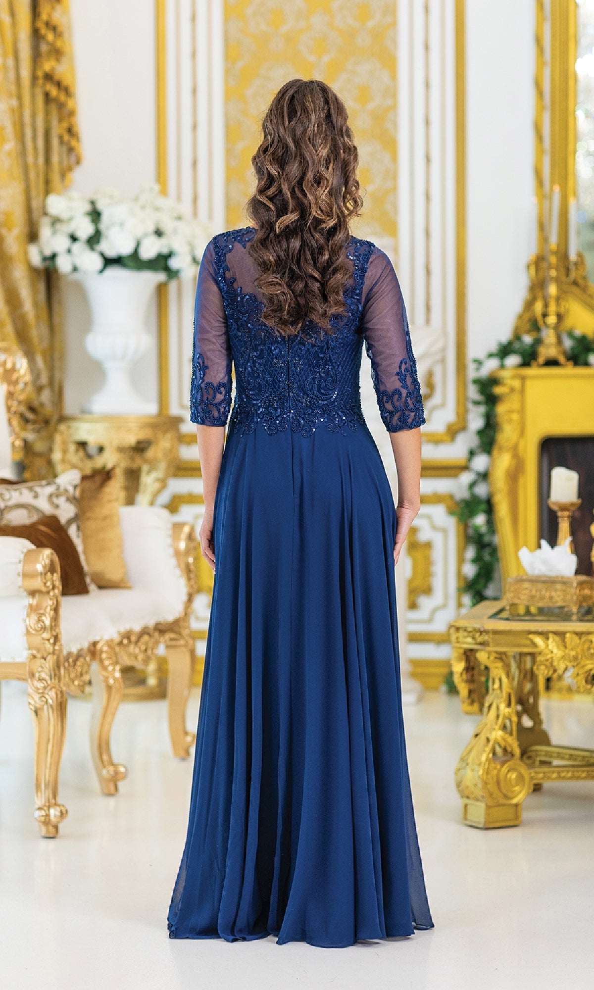 A woman with long curly hair stands facing away, wearing the Dancing Queen 4497 long sleeve formal dress with lace details and a beaded bodice, in an ornate room decorated in gold and white.