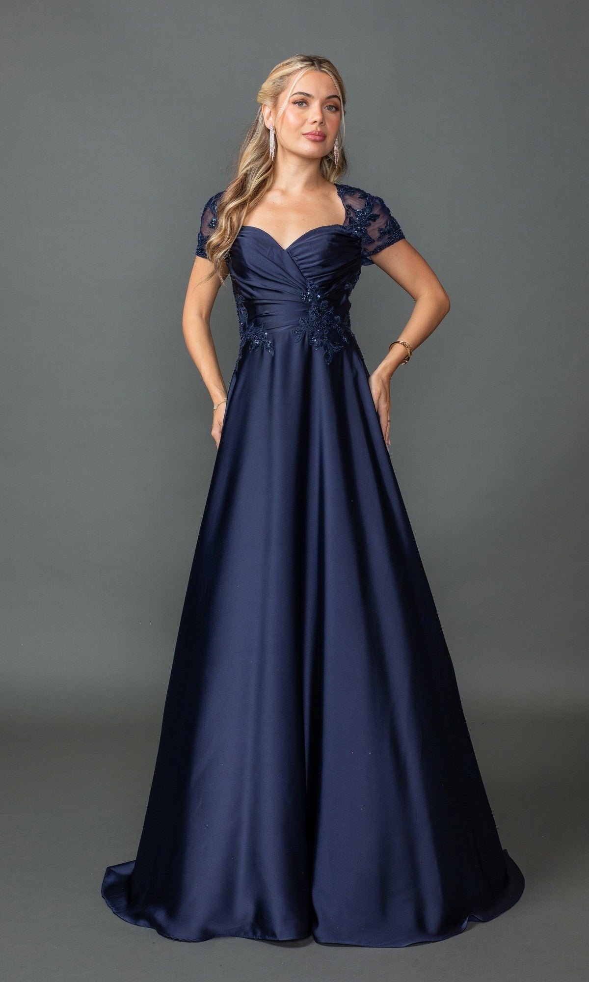 A woman stands against a plain background wearing the Dancing Queen 4499 short-sleeve long prom dress in navy, featuring lace sleeves and a fitted, ruched bodice.