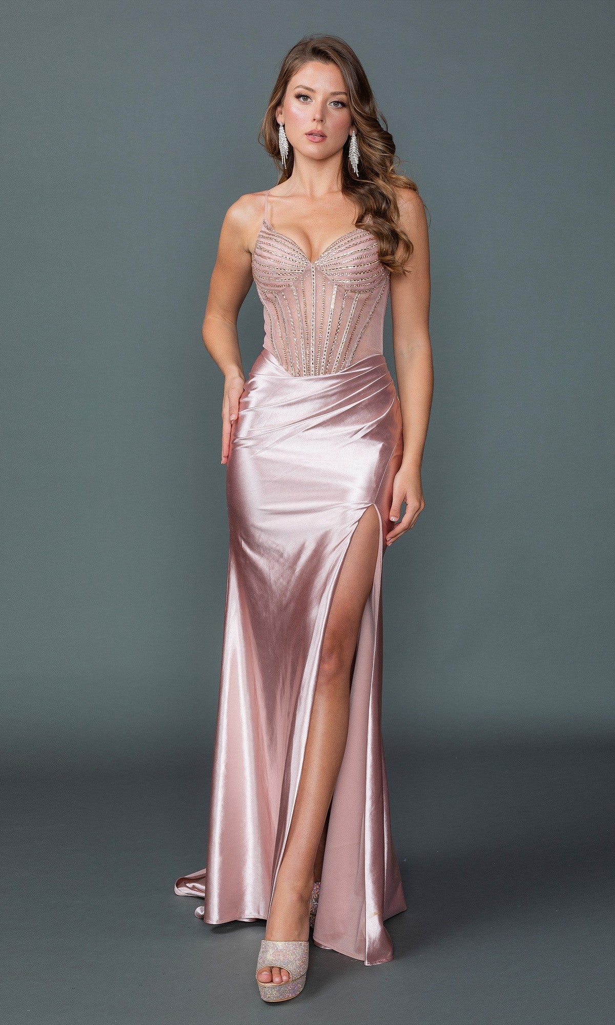 A woman poses against a plain gray background in the Sheer-Corset Long Prom Dress: Dancing Queen 4507—a fitted light pink satin gown with a sheer beaded bodice, spaghetti straps, and a high slit.
