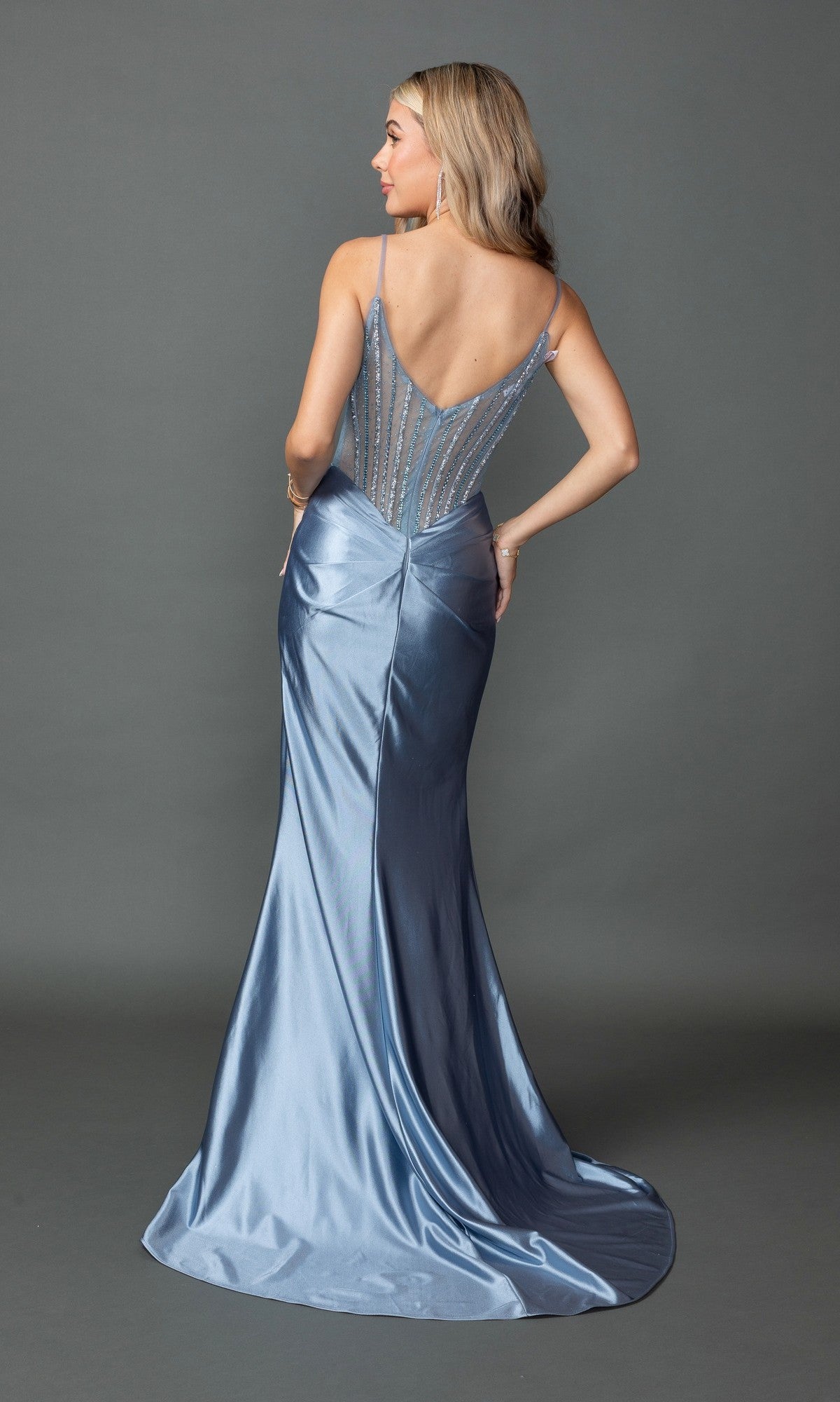 A woman poses with her back to the camera in a Sheer-Corset Long Prom Dress: Dancing Queen 4507, a floor-length light blue satin gown featuring a fitted, beaded bodice and thin straps, set against a gray backdrop.