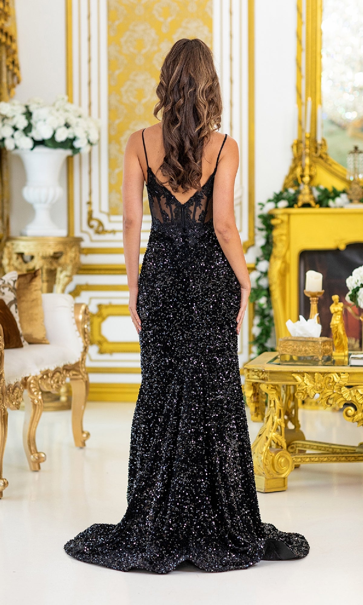 A woman in the Long Black Sequin Prom Dress: Dancing Queen 4511, featuring sequins and lace detailing on the back, stands facing away in an ornate, gold-accented room.