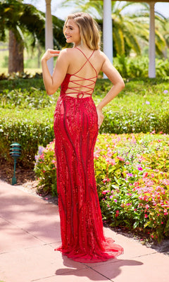 A woman wears the Primavera 4513 long red prom dress with beaded appliques, standing on a garden walkway surrounded by colorful flowers and greenery, smiling over her shoulder.