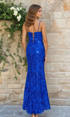A woman stands with her back to the camera in the Sequin-Lace Long Lace-Up Prom Dress 4521BN—a fitted, floor-length blue sequin gown with thin straps and lace-up detail—by a stone wall decorated with greenery and white flowers.