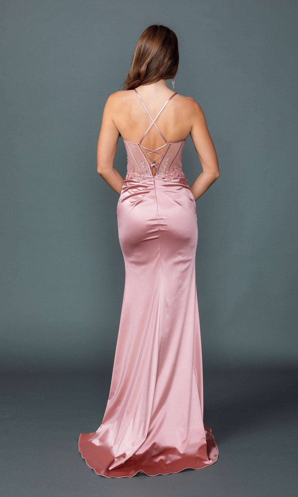 A woman in the Lace-Up Long Black Prom Dress: Dancing Queen 4524, with a lace-up back and thin straps, stands facing away to showcase the elegant black satin skirt against a plain gray background.