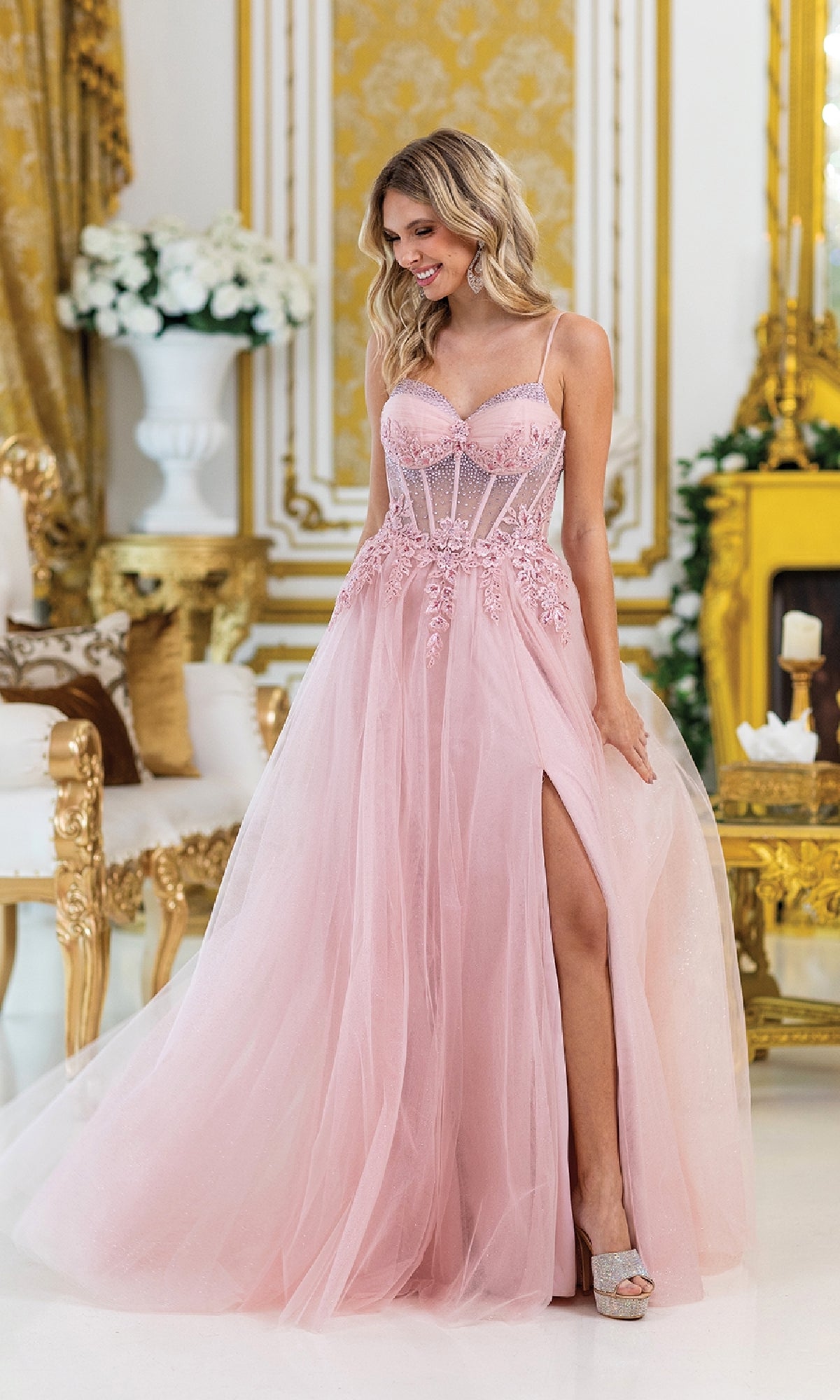A woman stands indoors in the Corset Prom Ball Gown with Slit: Dancing Queen 4529, a pink, sheer evening gown with lace embroidery and a high slit, surrounded by ornate gold and white decor.