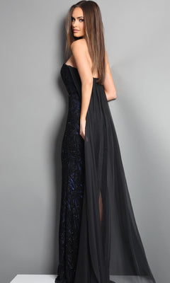A woman stands on a white platform against a gray background, looking over her shoulder while wearing the Jovani 45306 long formal dress—a strapless black evening gown with a sheer overlay. This elegant designer piece showcases sophisticated detailing and modern style, making it ideal for any upscale event.