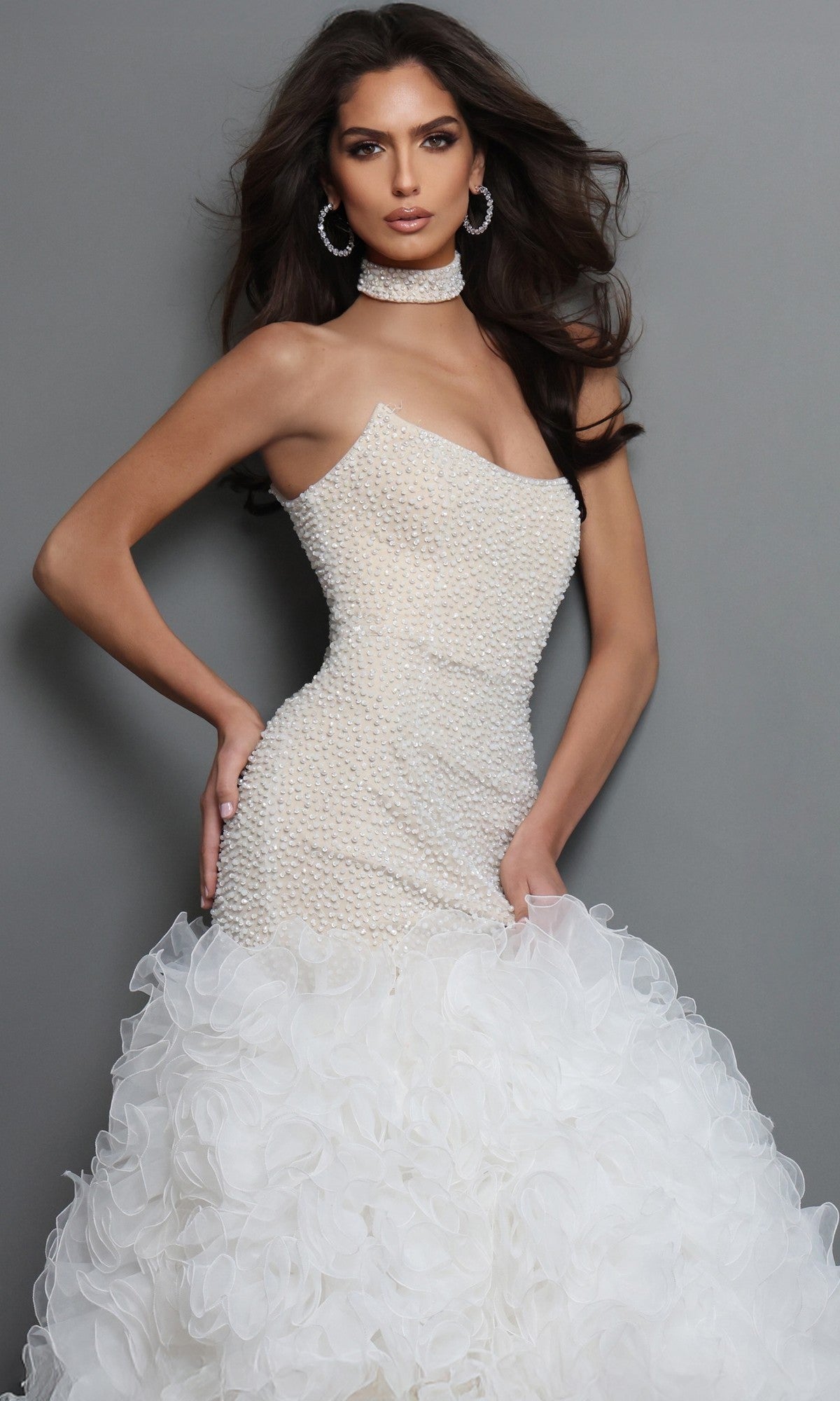 A woman poses in the Jovani 45339 Long Formal Dress, a white strapless designer gown featuring a fitted beaded bodice and a voluminous mermaid skirt with elegant ruffles, set against a gray background.