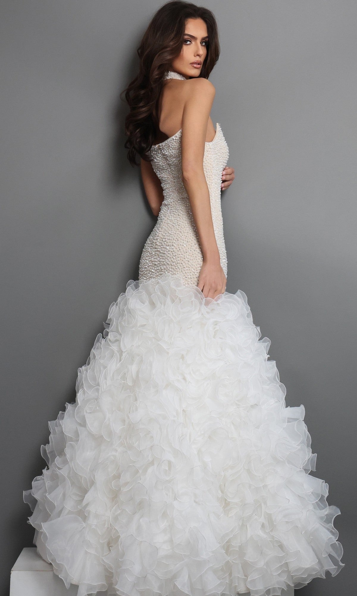 A woman stands against a gray background, looking over her shoulder, wearing the Long Formal Dress: Jovani 45339—a sleeveless, form-fitting designer gown that showcases a textured bodice and a striking mermaid skirt with delicate ruffled layers.