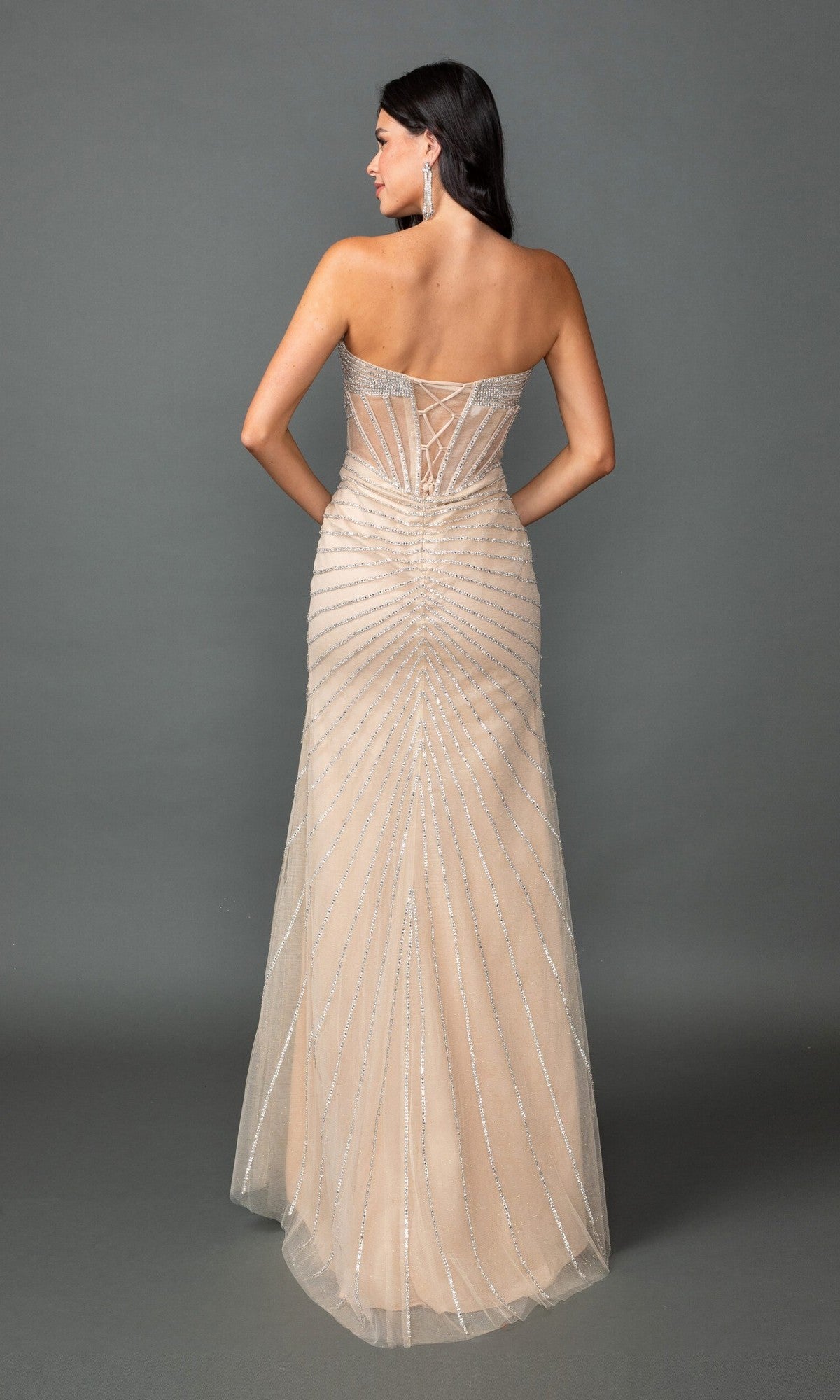 A woman stands against a plain gray background in the Dancing Queen 4562 long prom dress—a strapless, floor-length nude gown with a corset bodice, silver embellishments, and lace-up back detail.