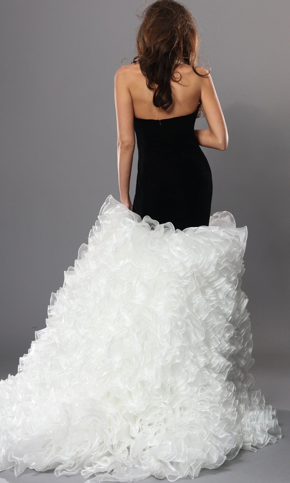 A woman with wavy brown hair faces away, showcasing the Jovani 45670 Long Formal Dress—a strapless gown with a velvet fitted bodice and a voluminous, white, ruffled tulle skirt that cascades into an elegant train—all set against a plain gray background.