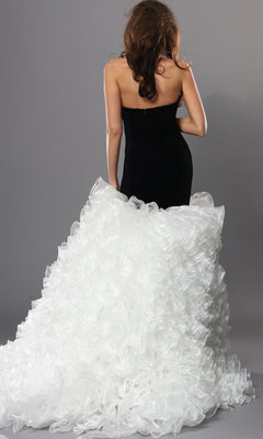 A woman with wavy brown hair faces away, showcasing the Jovani 45670 Long Formal Dress—a strapless gown with a velvet fitted bodice and a voluminous, white, ruffled tulle skirt that cascades into an elegant train—all set against a plain gray background.