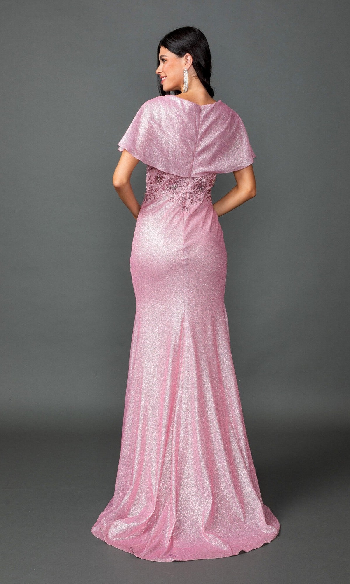 A woman stands facing away against a plain gray background, wearing the Dancing Queen 4568 long prom dress featuring shimmering pink fabric with cape sleeves and lace detailing.