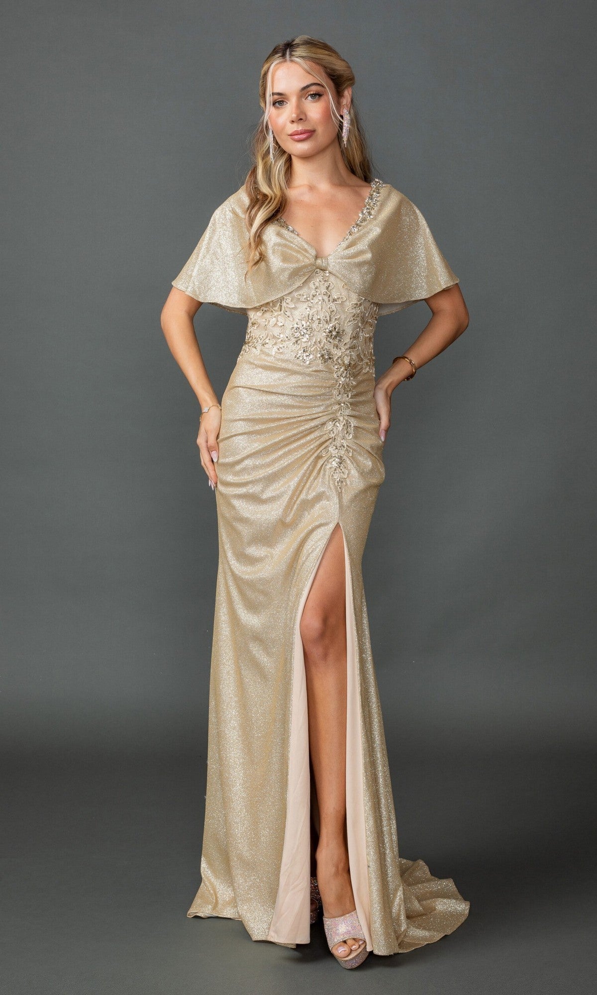 Wearing the Dancing Queen 4568, a champagne-colored long prom dress with cape sleeves and a side slit, a woman stands against a gray background in heels and minimal jewelry. The gown’s sheath silhouette enhances her elegant appearance.