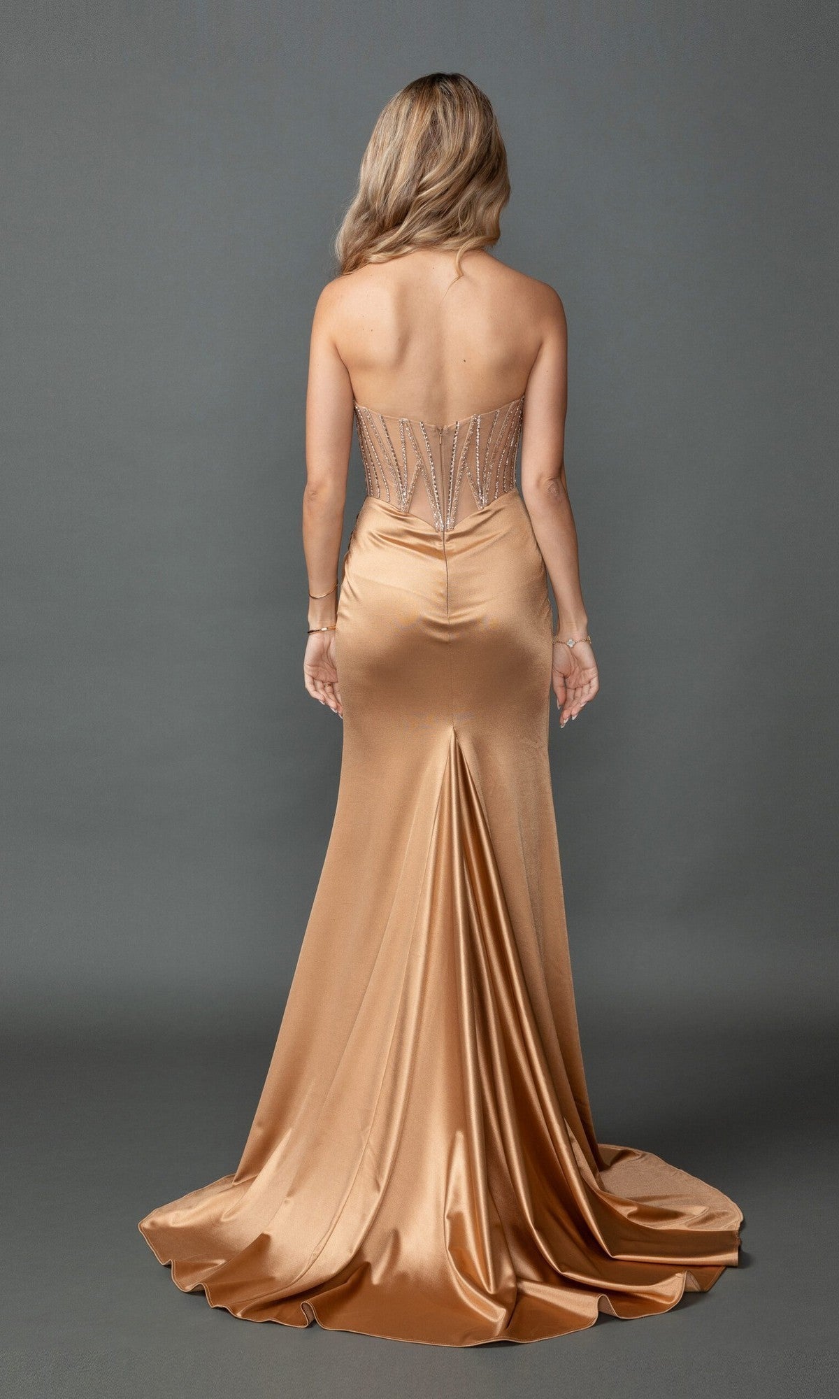 A woman stands facing away against a plain gray background, wearing the Long Prom Dress: Dancing Queen 4575—a gold strapless prom dress with a fitted, beaded corset bodice and a flared, floor-length satin sheath skirt.
