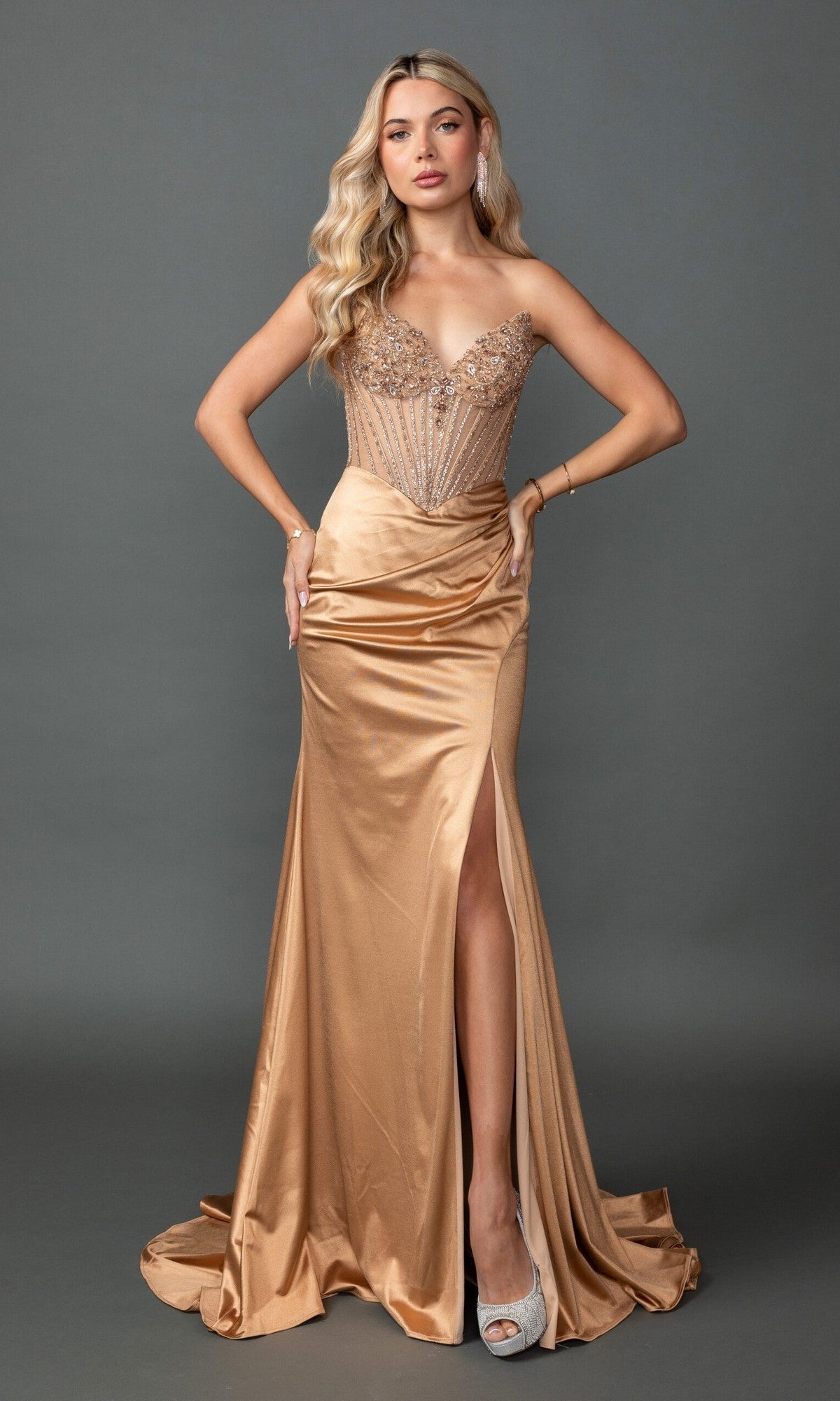 A woman wearing the Dancing Queen 4575 long gold prom dress—featuring a beaded corset bodice, satin sheath skirt with a thigh-high slit—paired with sparkling silver high heels, stands against a plain gray background.