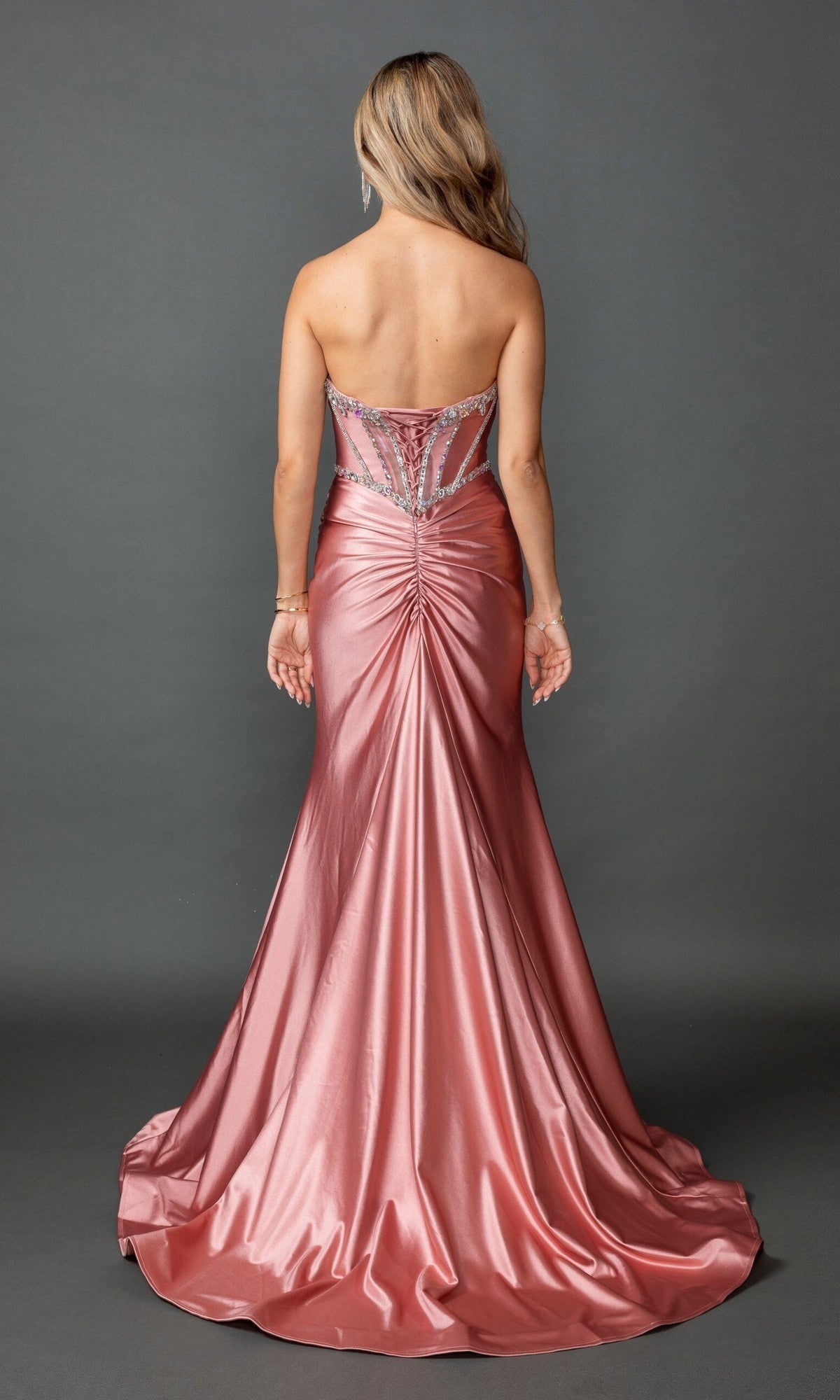 A woman stands facing away, wearing the Long Prom Dress: Dancing Queen 4580—a strapless metallic pink gown with a sleek corset bodice adorned with crystal bead embellishments and jeweled details. The dress features a beautifully gathered train and is set against a plain gray background.