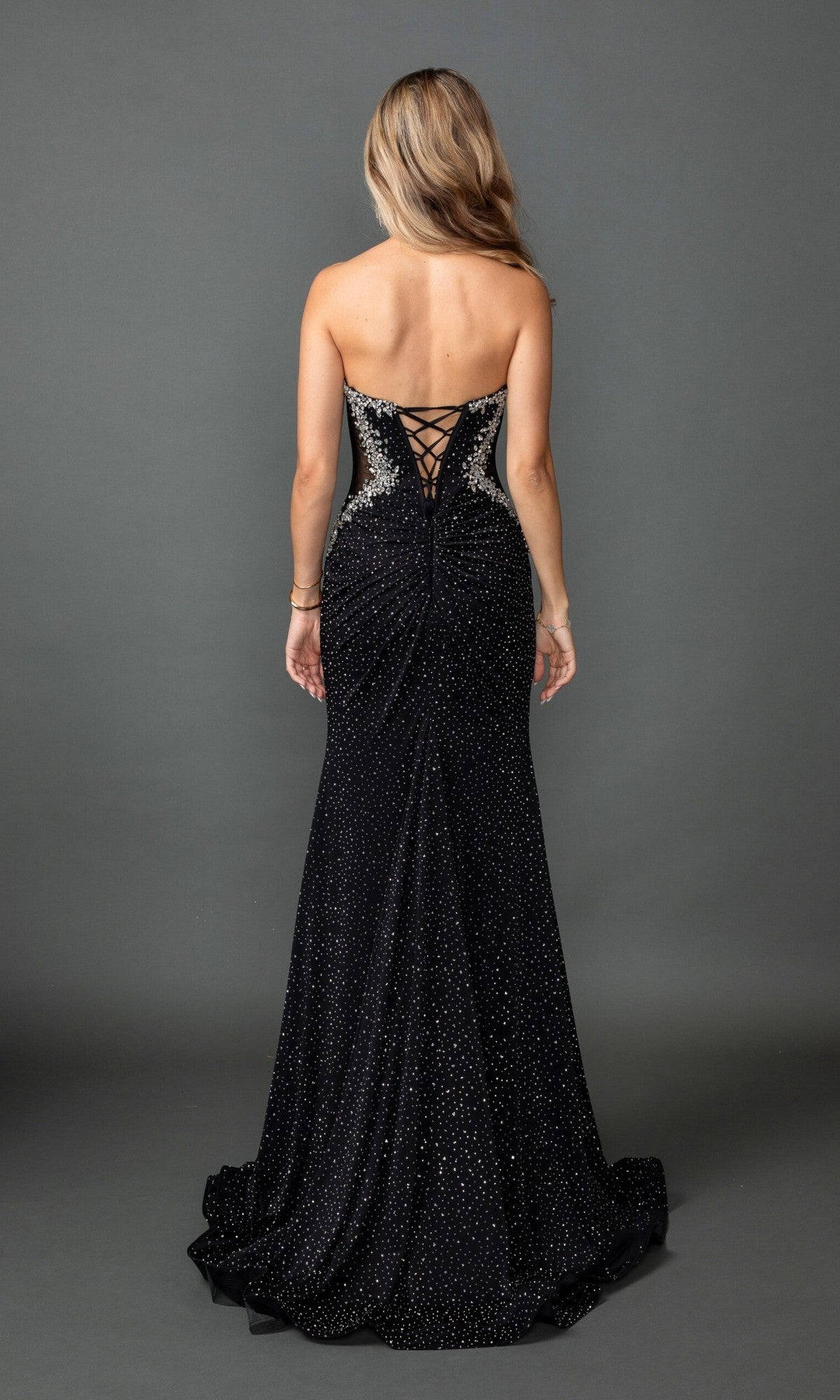 A woman with long, wavy hair is wearing the Dancing Queen 4583 long prom dress—a floor-length black gown featuring sparkly embellishments and a lace-up corset back—as she stands against a plain gray background.