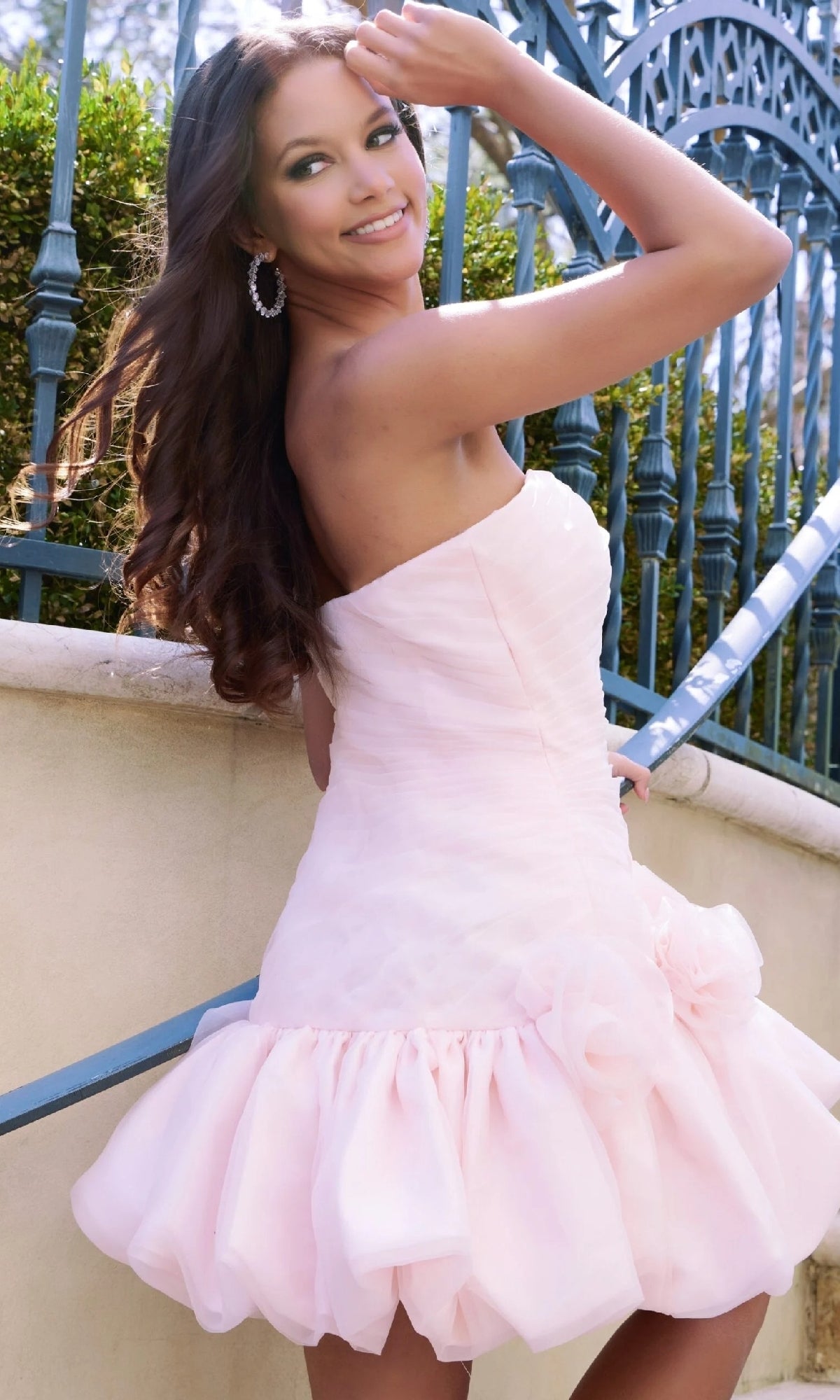 A woman stands outdoors by an ornate blue railing, smiling and holding her hair back while wearing the Jovani 45877 short homecoming dress in pale pink ruffles.